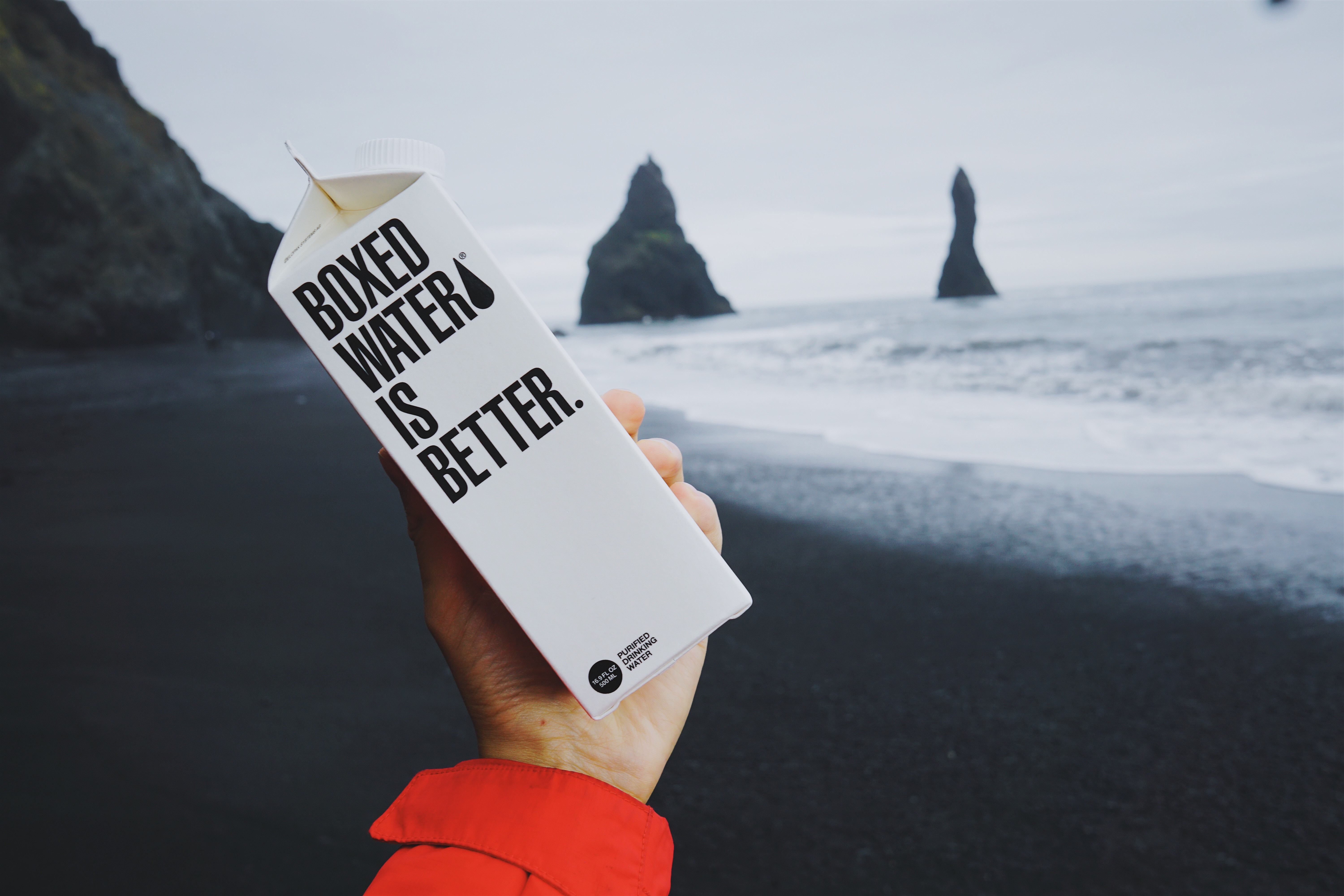 Arm holding up a carton that says 'boxed water is better'. Background of edge of ocean.